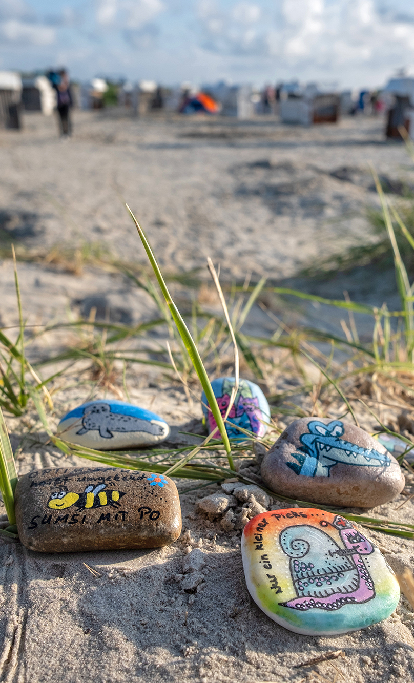 Bemalte Kieselsteine liegen am Strand an der Nordseeküste. Im Hintergrund sind Personen und Strandkörbe zu sehen.