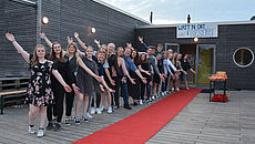 Eine Gruppe junger Menschen steht begrüßend vor einem roten Teppich der zum Eingang eines Gebäudes führt.
