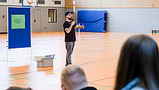 Dominik Forster steht in einer Turnhalle und spricht zu einem Publikum von Jugendlichen