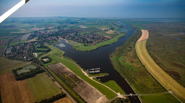 Luftaufnahme einer Küstenlandschaft mit einem Flusslauf, Deichen und einem Ort mit roten Dächern, umgeben von Feldern, Wiesen und weitläufigem Marschland.