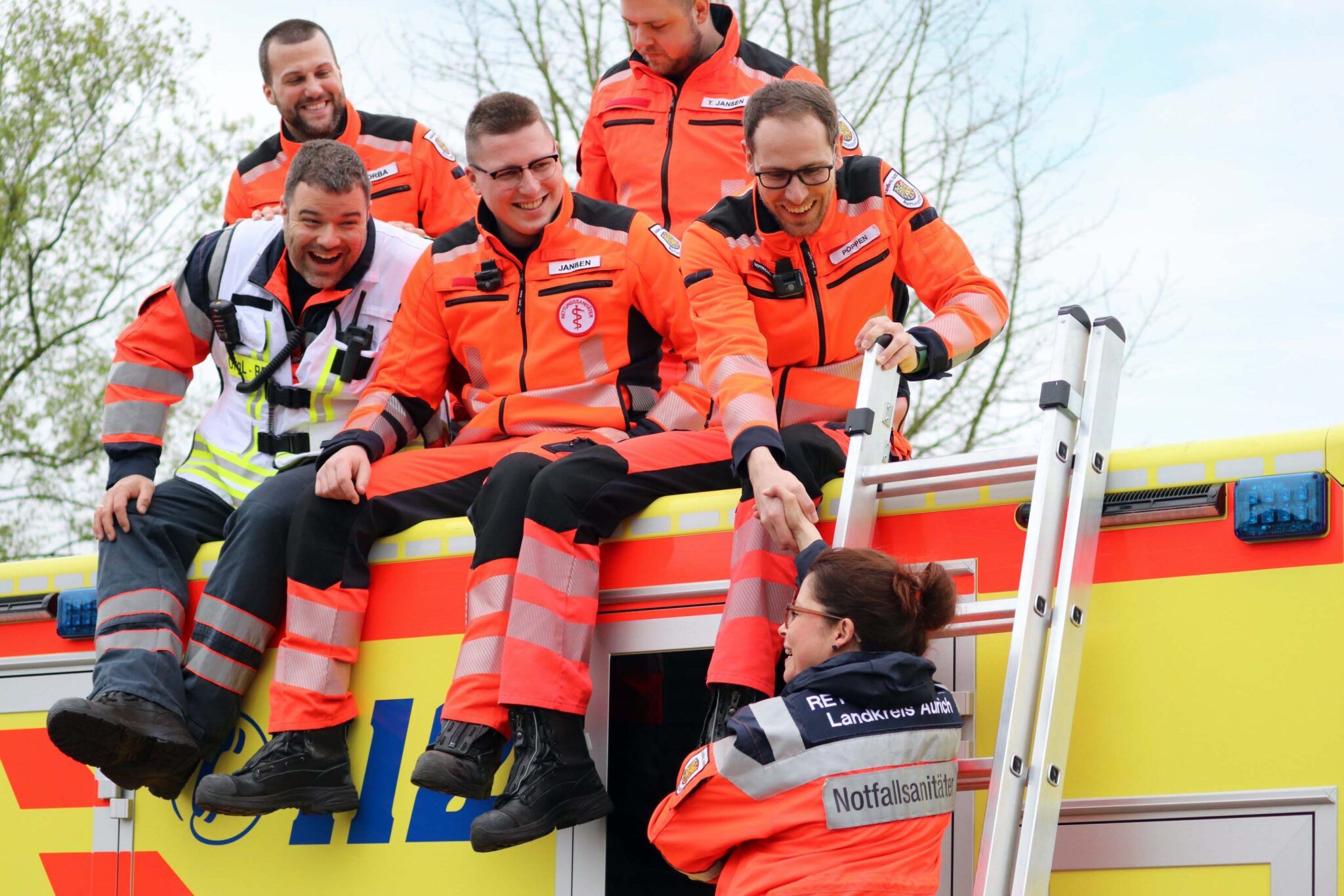 Mehrere Rettungskräfte in orangefarbener Einsatzkleidung sitzen lachend auf dem Dach eines Rettungswagens. Eine Kollegin in Uniform mit der Aufschrift ‚Rettungsdienst Landkreis Aurich‘ und ‚Notfallsanitäterin‘ steht auf einer Leiter und reicht einem Kollegen die Hand. Die Szene wirkt freundschaftlich und kollegial.