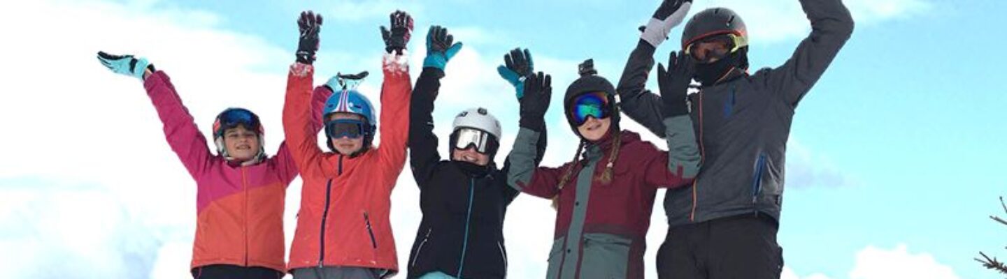 Fünf Kinder in Winterkleidung stehen im Schnee und strecken jubelnd die Arme in die Höhe, vor blauem Himmel.