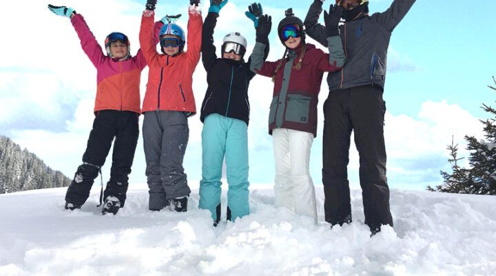 Fünf Kinder in Winterkleidung stehen im Schnee und strecken jubelnd die Arme in die Höhe, vor blauem Himmel.