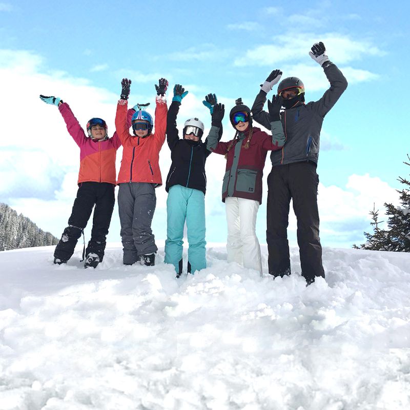 Fünf Kinder in Winterkleidung stehen im Schnee und strecken jubelnd die Arme in die Höhe, vor blauem Himmel.