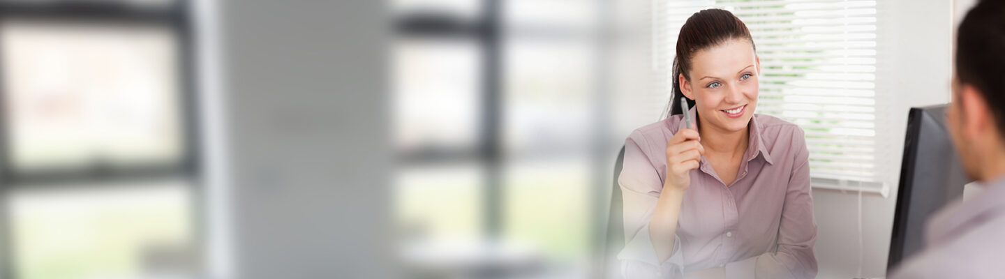 Frau in einem Bürogespräch: Eine Mitarbeiterin sitzt an einem Schreibtisch, hält einen Stift in der Hand und spricht freundlich mit einer Person gegenüber, die nur teilweise sichtbar ist. Im Hintergrund sind Fenster mit Jalousien zu sehen.
