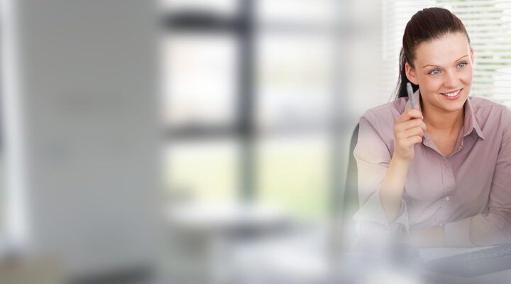 Frau in einem Bürogespräch: Eine Mitarbeiterin sitzt an einem Schreibtisch, hält einen Stift in der Hand und spricht freundlich mit einer Person gegenüber, die nur teilweise sichtbar ist. Im Hintergrund sind Fenster mit Jalousien zu sehen.