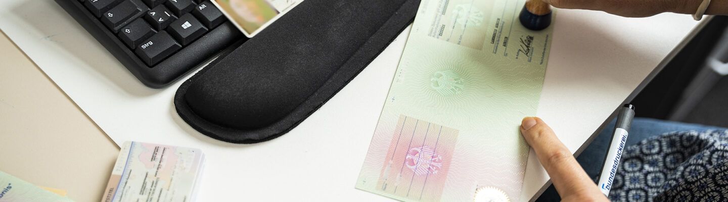 Eine Person stempelt ein amtliches Dokument auf einem Schreibtisch. Daneben liegen eine Tastatur, ein Ausweisdokument sowie weitere Unterlagen.