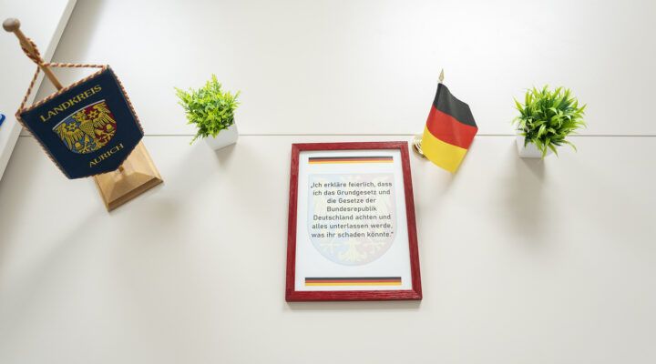 An einer Wand hängen eine kleine Fahne mit dem Wappen des Landkreises Aurich, zwei grüne Pflanzen in weißen Töpfen und eine Deutschlandflagge. In der Mitte befindet sich ein gerahmtes Dokument mit dem Amtseid auf das Grundgesetz der Bundesrepublik Deutschland.