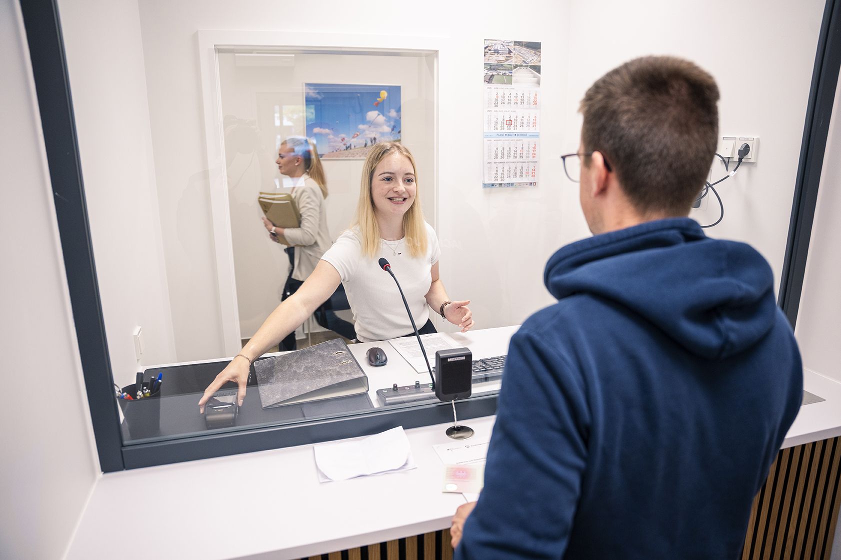 Eine Mitarbeiterin sitzt hinter einer Glasscheibe an einem Serviceschalter und spricht freundlich mit einem Mann auf der anderen Seite. Auf dem Tresen liegen Unterlagen, ein Mikrofon und ein Stempel; im Hintergrund ist eine weitere Mitarbeiterin mit Aktenordnern zu sehen.