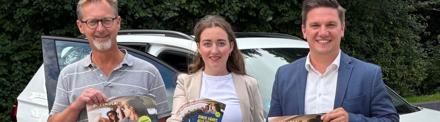 Ingo de Vries (Klimaschutzmanagement), Sonja Wolke und Geschäftsführer Jelto Müller (Wirtschaftsförderung Landkreis Aurich GmbH) stehen vor einem Auto und halten Werbeplakate für das Angebot "PENDLER"