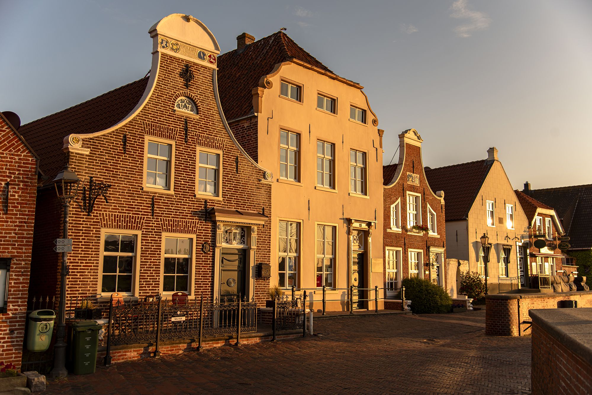 Historische Backsteinhäuser in Greetsiel, mit geschwungenen Giebeln stehen in einer Reihe entlang einer gepflasterten Straße. Die Fassaden sind von warmem Abendlicht angestrahlt. Vor den Häusern befinden sich Fenster mit weißen Rahmen, verzierte Türen, schmiedeeiserne Zäune und klassische Straßenlaternen.