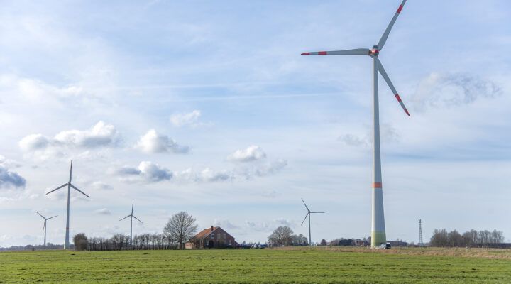 Weite Landschaft mit grünen Wiesen, mehreren Windkraftanlagen und einem einzelnen Bauernhaus in der Mitte. Der Himmel ist leicht bewölkt, die Windräder prägen die ländliche Umgebung.