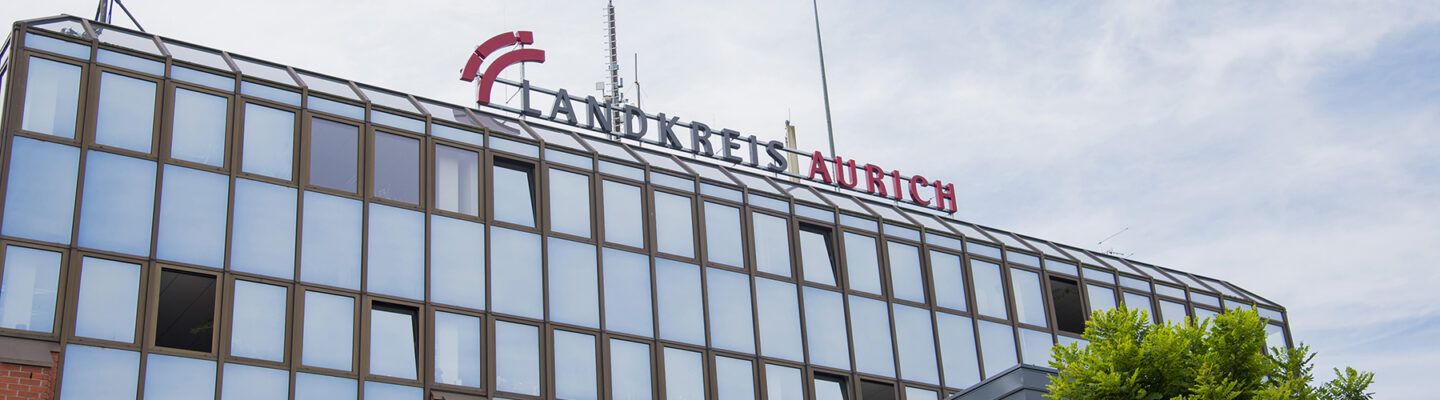 Außenansicht eines Verwaltungsgebäudes mit Glasfassade. Auf dem Dach ist der Schriftzug ‚Landkreis Aurich‘ angebracht; darüber ragt eine Antennenanlage in den Himmel.