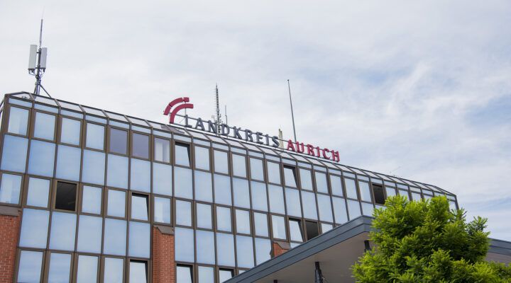 Außenansicht eines Verwaltungsgebäudes mit Glasfassade. Auf dem Dach ist der Schriftzug ‚Landkreis Aurich‘ angebracht; darüber ragt eine Antennenanlage in den Himmel.