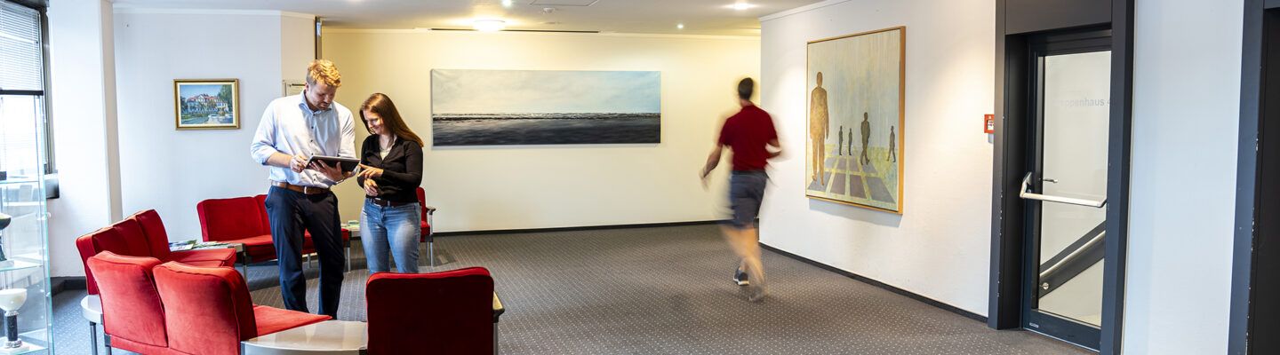 Heller Flur mit Sitzbereich und Kunst an den Wänden. Zwei Personen stehen zusammen und schauen auf ein Tablet, während eine weitere Person im Hintergrund durch den Flur geht.