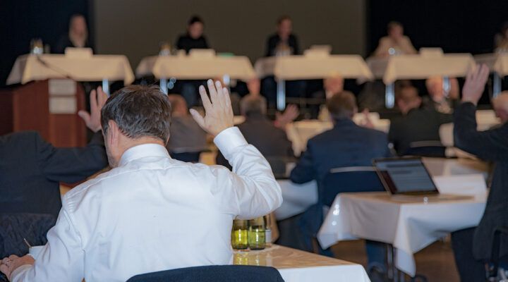 Mehrere Personen sitzen in einem Konferenzraum an Tischen; im Vordergrund hebt ein Mann im weißen Hemd die Hand, auch weitere Teilnehmende melden sich, während vorne eine Sitzung stattfindet.