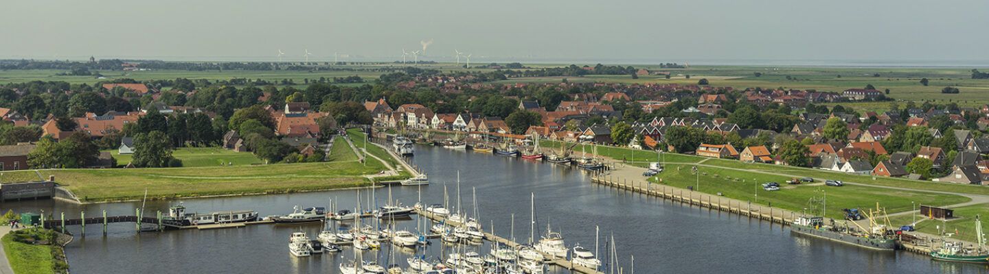 Luftaufnahme eines kleinen Küstenortes mit Hafenbecken und zahlreichen Segelbooten. Dahinter liegen Häuser mit roten Ziegeldächern, grüne Deichflächen und weite Marschlandschaft bis zum Wattenmeer; am Horizont sind mehrere Windkraftanlagen zu sehen.