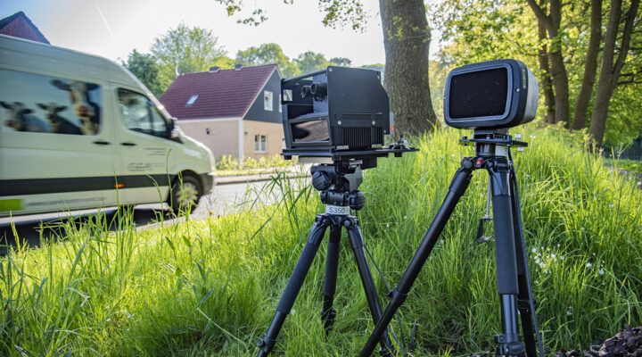 Mobiles Geschwindigkeitsmessgerät (Blitzer) am Straßenrand auf einem Stativ aufgebaut. Im Hintergrund fährt ein heller Lieferwagen an einem Wohnhaus vorbei. Die Szene befindet sich in einem grünen Wohngebiet mit Bäumen und hohem Gras.