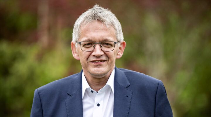 Porträt von Landrat Olaf Meinen. Er trägt ein weißes Hemd und ein dunkelblaues Sakko und steht vor einem unscharfen, grünlichen Hintergrund im Freien.