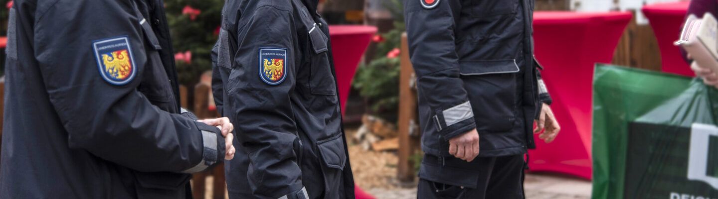 Mehrere Einsatzkräfte des Landkreises Aurich stehen nebeneinander bei einer Veranstaltung im Freien. Auf ihren dunklen Jacken ist das Wappen des Landkreises Aurich zu sehen. Im Hintergrund befinden sich Stehtische mit roten Hussen.