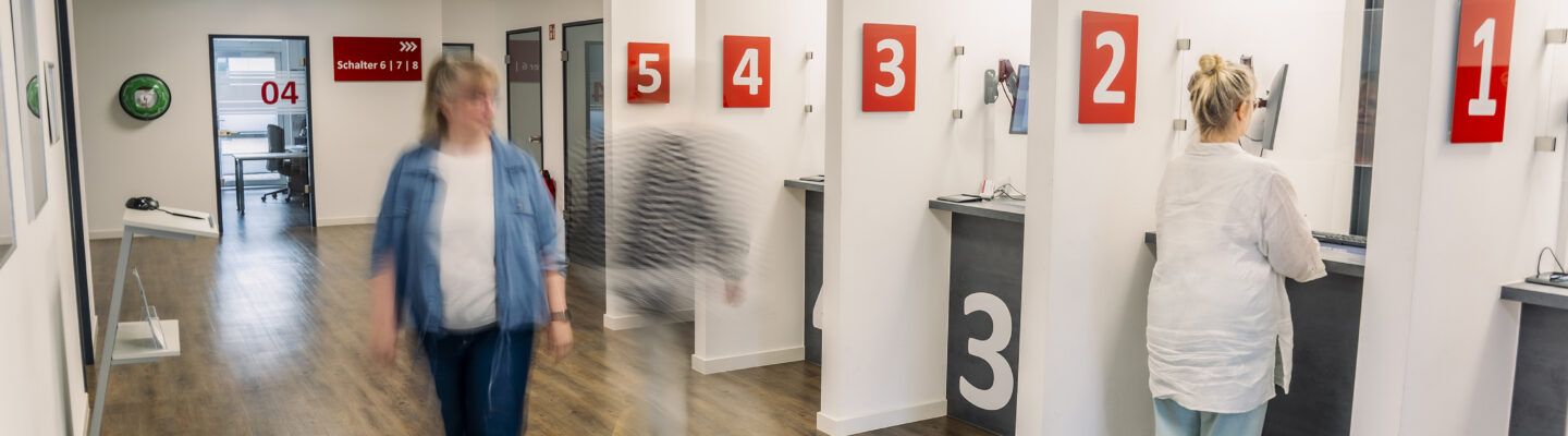 Bürgerbüro mit mehreren nummerierten Serviceschaltern (1–5) an einer hellen Wand. Eine Mitarbeiterin bedient eine Person am Schalter, während andere Personen unscharf durch den Flur gehen. Im Hintergrund sind weitere Räume und ein Hinweisschild zu Schalter 6–8 sichtbar.