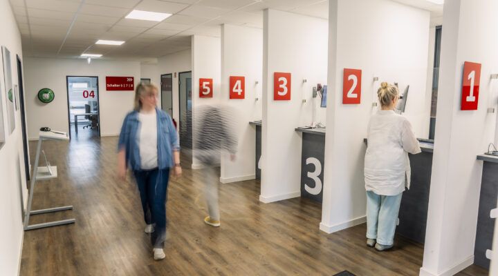 Bürgerbüro mit mehreren nummerierten Serviceschaltern (1–5) an einer hellen Wand. Eine Mitarbeiterin bedient eine Person am Schalter, während andere Personen unscharf durch den Flur gehen. Im Hintergrund sind weitere Räume und ein Hinweisschild zu Schalter 6–8 sichtbar.