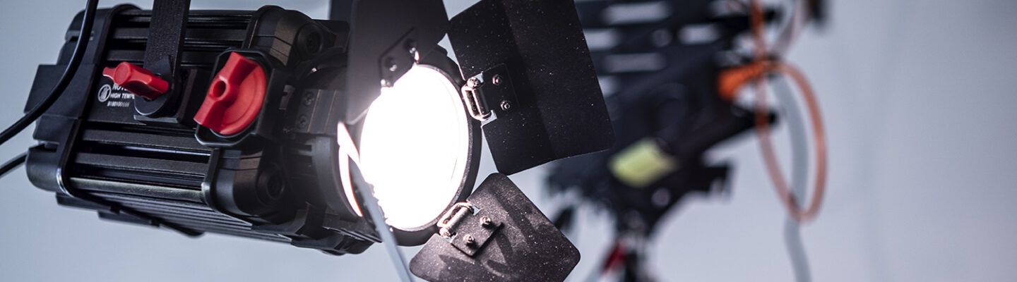 Ein eingeschalteter Studioscheinwerfer mit Flügeltoren leuchtet hell in einem Innenraum. Im Hintergrund ist weiteres Beleuchtungsequipment unscharf zu erkennen.