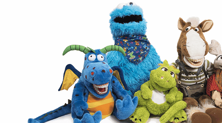 Mehrere bunte Handpuppen sitzen nebeneinander vor weißem Hintergrund. Zu sehen sind unter anderem ein blauer Drache, ein grünes Tier, ein Pferd, eine Maus, ein Frosch, ein weißer Clown sowie eine große blaue Figur im Hintergrund.