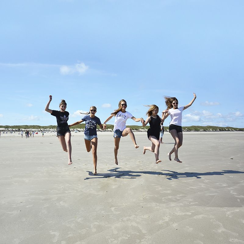 Fünf Jugendliche springen lachend barfuß am Strand in die Luft, bei sonnigem Wetter und blauem Himmel.