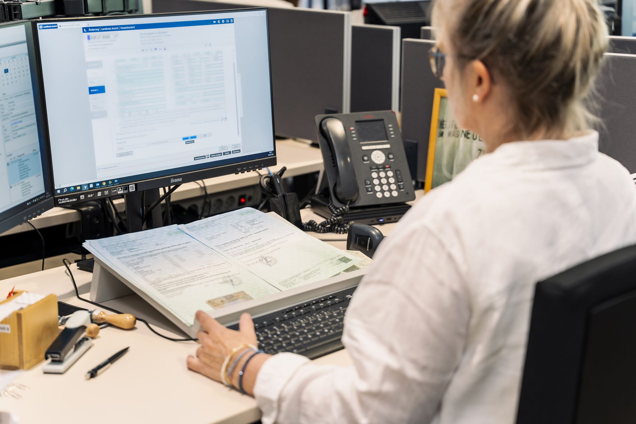 Eine Mitarbeiterin sitzt an einem Büroarbeitsplatz vor zwei Monitoren. Auf dem Schreibtisch liegen geöffnete Dokumente, ein Stempel sowie weitere Büroutensilien. Auf dem Bildschirm ist eine digitale Verwaltungsoberfläche zu sehen, während sie Unterlagen bearbeitet.