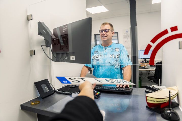Mitarbeiter hinter einer Glasscheibe am Serviceschalter, trägt ein blaues T-Shirt und spricht mit einer Person auf der anderen Seite. Vor ihm stehen Monitor, Tastatur und Kartenlesegerät; auf dem Tresen liegt ein Ausweis oder Dokument.