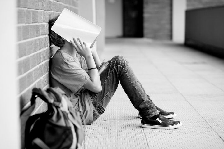 Schwarz-weiß-Foto eines Kindes, das auf dem Boden an eine Backsteinwand gelehnt sitzt und ein Buch vor das Gesicht hält; neben ihm liegt ein Rucksack in einem Flur oder Gang.