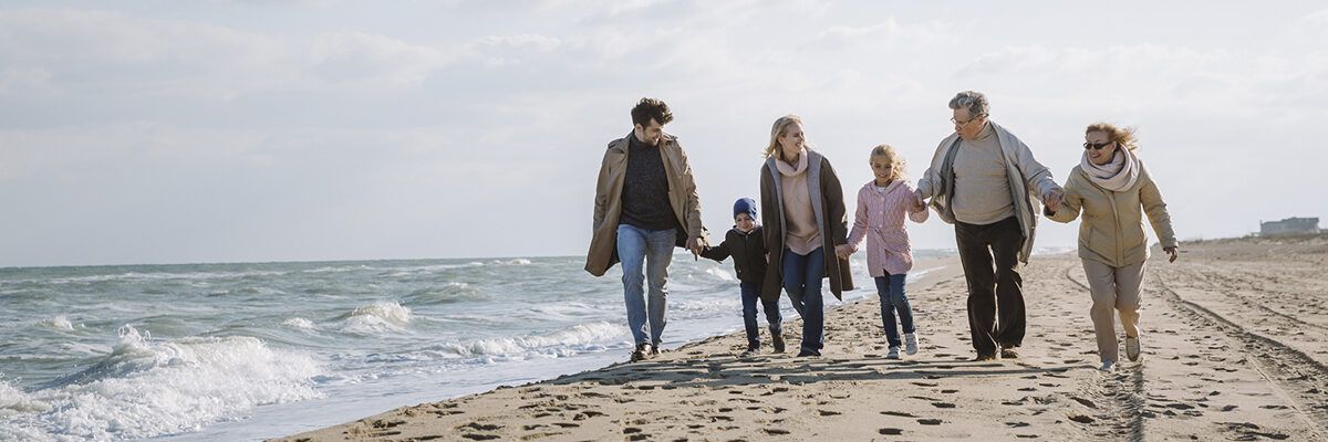 Mehrgenerationen-Familie spaziert Hand in Hand am Strand entlang; links das Meer mit Wellen, rechts der Sandstrand, helles Tageslicht und offener Himmel.