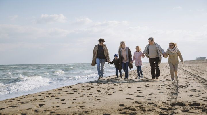 Mehrgenerationen-Familie spaziert Hand in Hand am Strand entlang; links das Meer mit Wellen, rechts der Sandstrand, helles Tageslicht und offener Himmel.