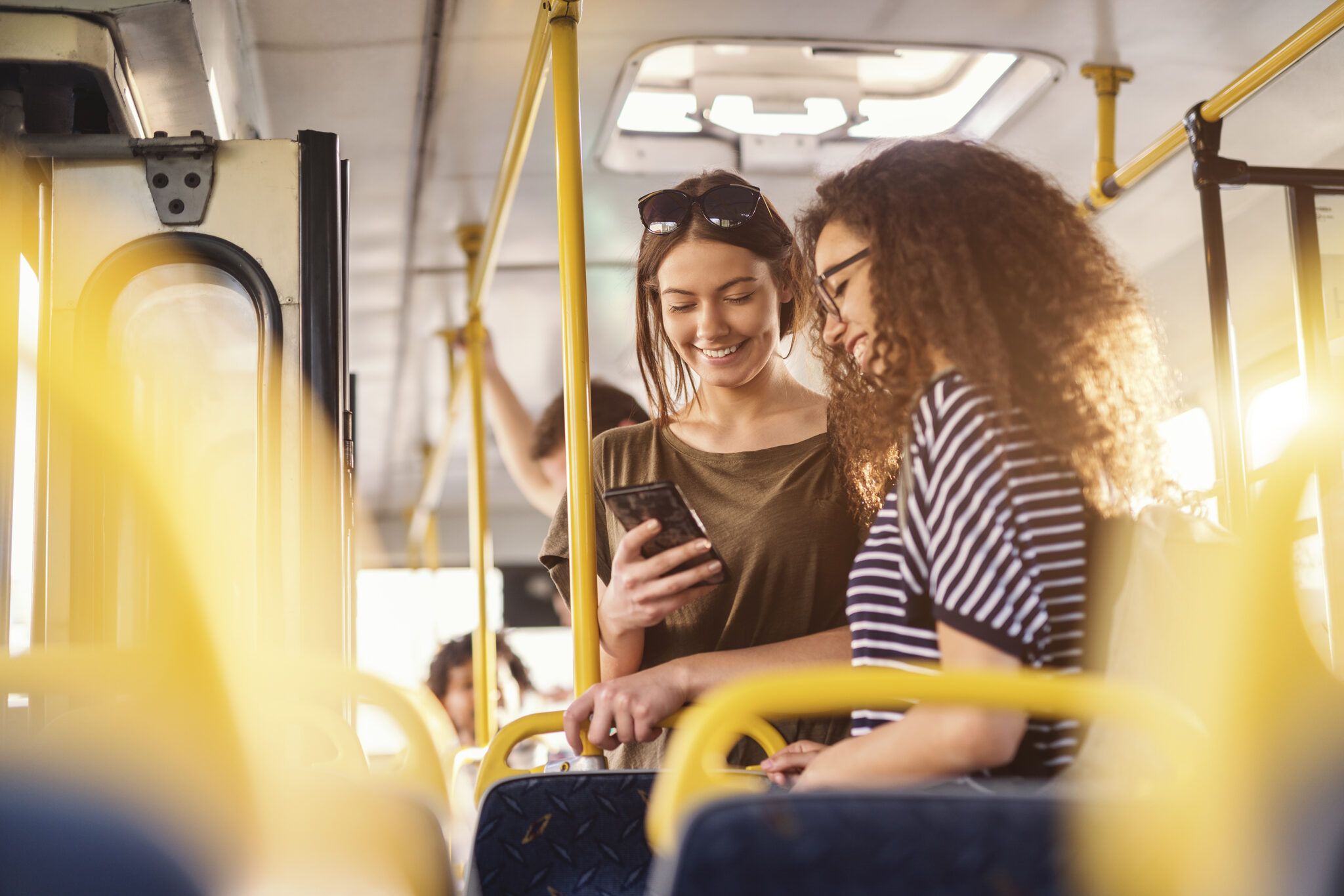 Zwei junge Frauen stehen in einem Linienbus nebeneinander und schauen lächelnd auf ein Smartphone; sie halten sich an gelben Haltestangen fest, im Hintergrund sind weitere Fahrgäste zu sehen.