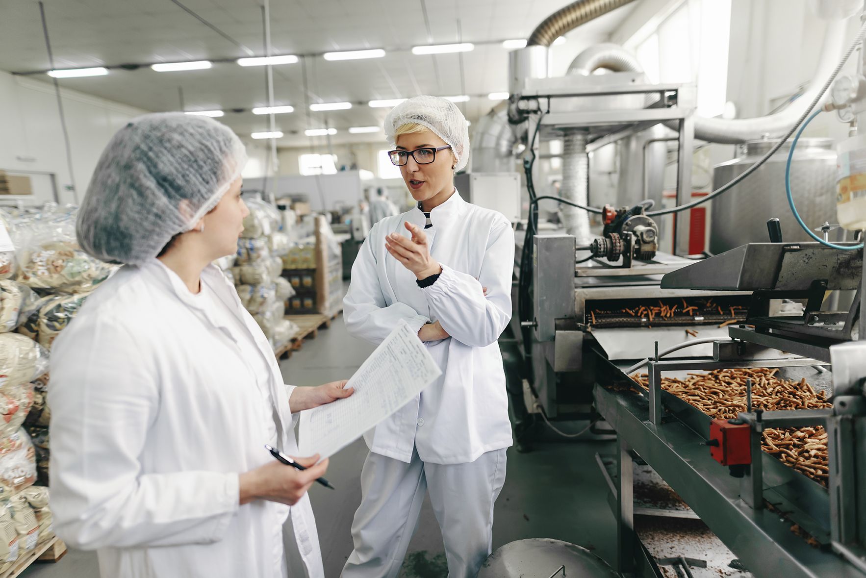 Lebensmittelkontrolleurin und Fabrikmitarbeiterin in weißen Uniformen und mit sterilen Hauben auf den Köpfen unterhalten sich über die Hygienequalität, während sie in einer Lebensmittelfabrik stehen.