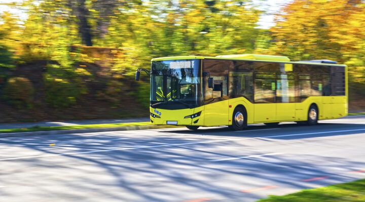 Gelber Linienbus fährt auf einer Straße entlang, umgeben von herbstlich gefärbten Bäumen; das Fahrzeug ist leicht verschwommen, was die Bewegung betont.
