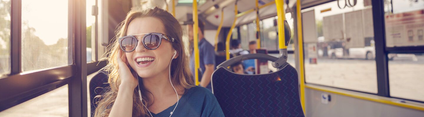 Junge Frau mit Sonnenbrille sitzt in einem Bus am Fenster, lächelt und hört mit Kopfhörern Musik; warmes Sonnenlicht fällt durch das Fenster ins Fahrzeug.