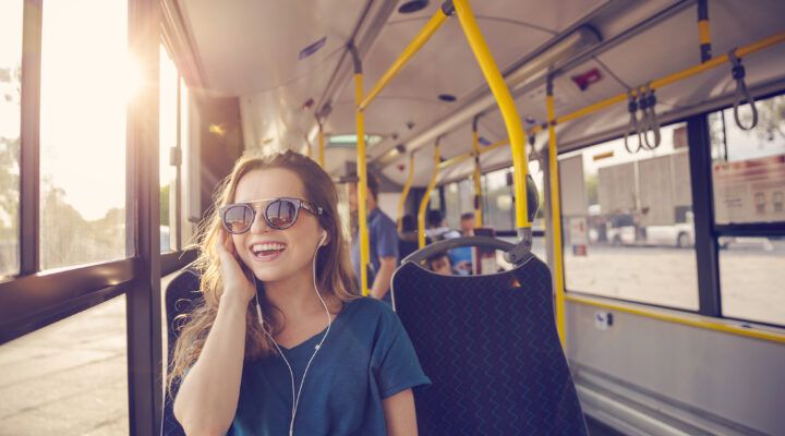 Junge Frau mit Sonnenbrille sitzt in einem Bus am Fenster, lächelt und hört mit Kopfhörern Musik; warmes Sonnenlicht fällt durch das Fenster ins Fahrzeug.