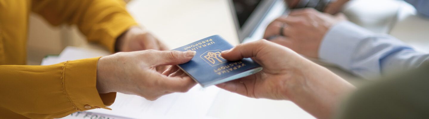 Eine Person übergibt einer anderen Person einen ukrainischen Reisepass über einen Schreibtisch hinweg. Im Hintergrund sind unscharf weitere Unterlagen und eine dritte Person zu erkennen.