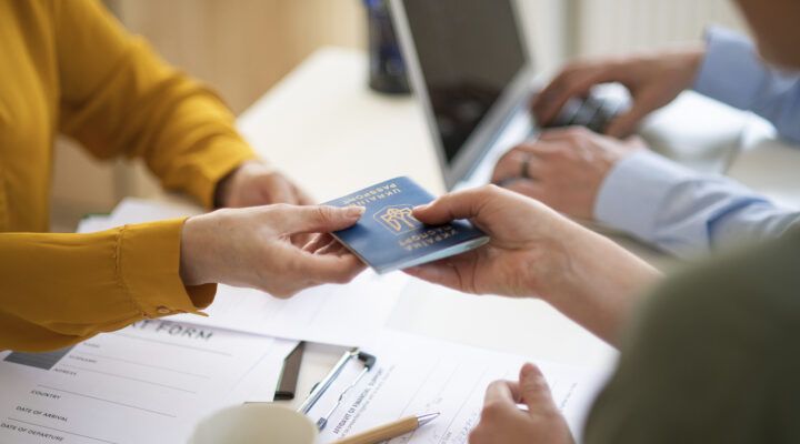 Eine Person übergibt einer anderen Person einen ukrainischen Reisepass über einen Schreibtisch hinweg. Im Hintergrund sind unscharf weitere Unterlagen und eine dritte Person zu erkennen.