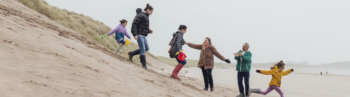 Mehrere Menschen unterschiedlichen Alters laufen und spielen gemeinsam an einem windigen Strand entlang einer Sanddüne; Kinder und Erwachsene bewegen sich lachend über den Sand.