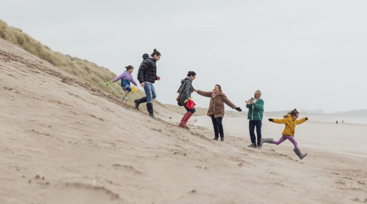 Mehrere Menschen unterschiedlichen Alters laufen und spielen gemeinsam an einem windigen Strand entlang einer Sanddüne; Kinder und Erwachsene bewegen sich lachend über den Sand.