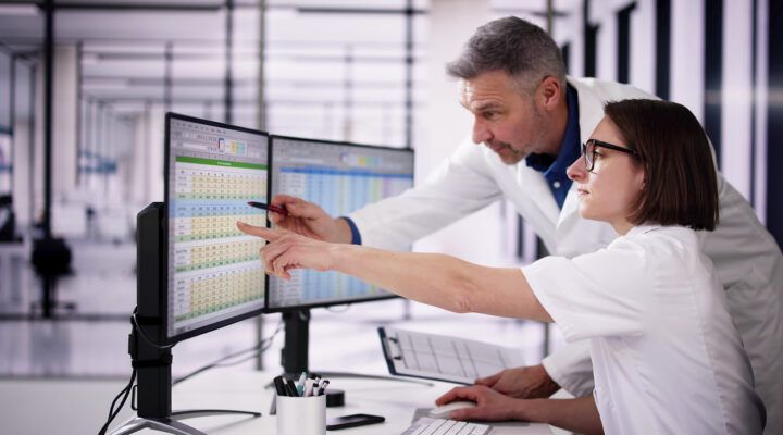 Zwei Personen in Laborkitteln arbeiten gemeinsam an einem Schreibtisch und betrachten Daten auf zwei Computerbildschirmen; eine Person zeigt mit dem Finger auf eine Tabellenansicht, während die andere mit einem Stift auf den Bildschirm weist. Im Hintergrund ist ein modernes Laborumfeld zu sehen.