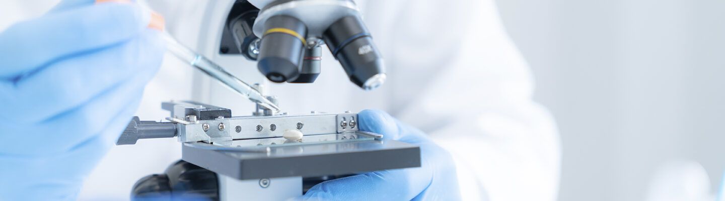 Nahaufnahme eines Labormikroskops. Eine Person mit blauen Einweghandschuhen platziert mit einer Pipette eine Probe auf einem Objektträger unter dem Mikroskop. Die Szene ist hell und klinisch ausgeleuchtet.