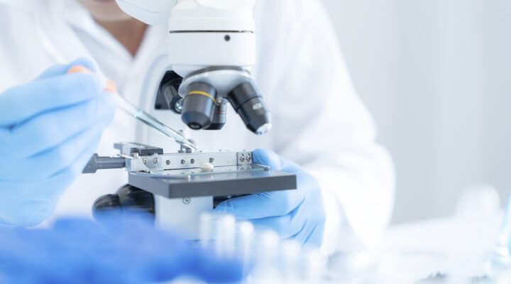 Nahaufnahme eines Labormikroskops. Eine Person mit blauen Einweghandschuhen platziert mit einer Pipette eine Probe auf einem Objektträger unter dem Mikroskop. Die Szene ist hell und klinisch ausgeleuchtet.