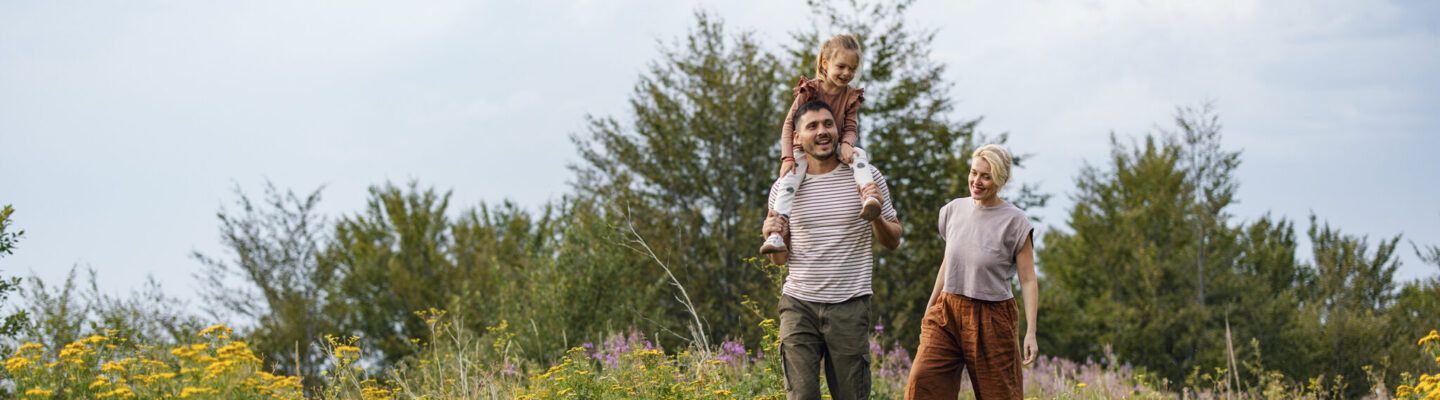 Eine Familie spaziert durch eine sommerliche Wiese mit Wildblumen. Ein Mann trägt ein kleines Kind auf den Schultern, beide lachen. Neben ihm geht eine Frau und lächelt die beiden an. Im Hintergrund stehen grüne Bäume unter einem leicht bewölkten Himmel.