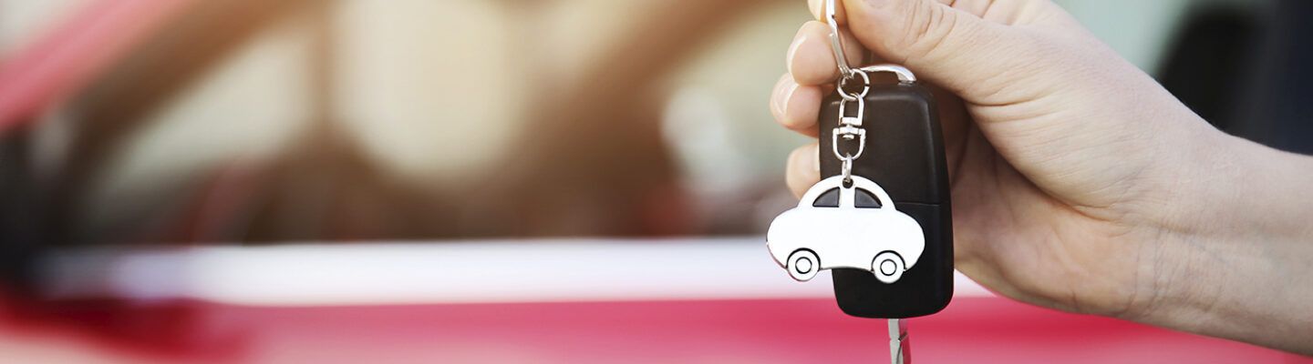 Hand hält einen Autoschlüssel mit schwarzem Funkschlüssel und einem weißen Anhänger in Form eines Autos; im Hintergrund ist unscharf ein rotes Fahrzeug zu sehen.