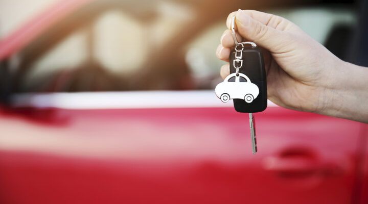 Hand hält einen Autoschlüssel mit schwarzem Funkschlüssel und einem weißen Anhänger in Form eines Autos; im Hintergrund ist unscharf ein rotes Fahrzeug zu sehen.