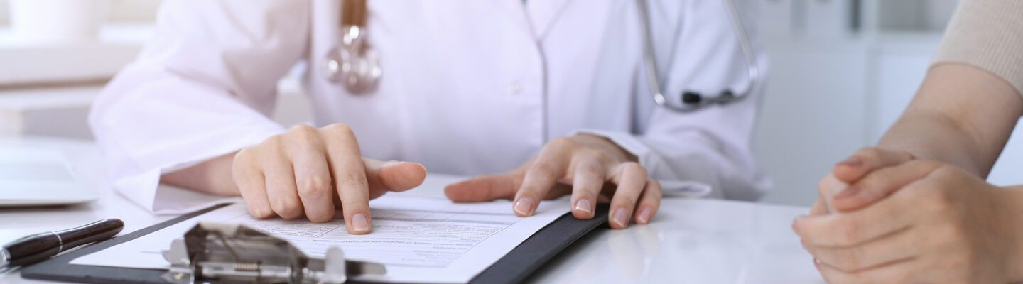 Ärztin oder Arzt im weißen Kittel erklärt einer Person ein Dokument auf einem Klemmbrett und zeigt mit dem Finger auf einen Abschnitt; gegenüber sitzen die gefalteten Hände der zu beratenden Person auf dem Tisch.